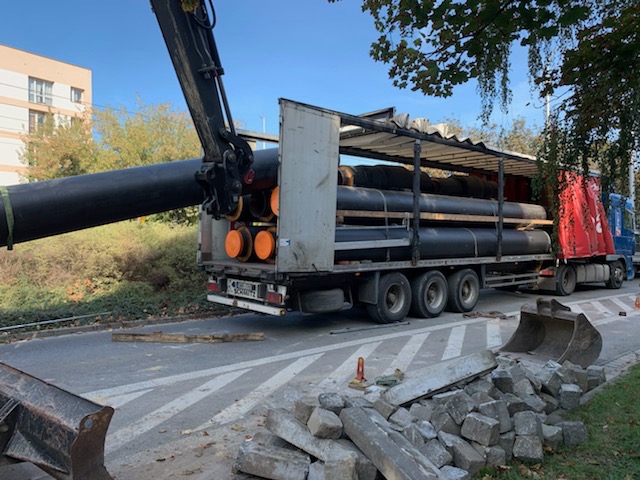 Ciężarówka z naczepą załadowana rurami, wyładowywana dźwigiem na placu budowy. Rury zabezpieczone pomarańczowymi zaślepkami.