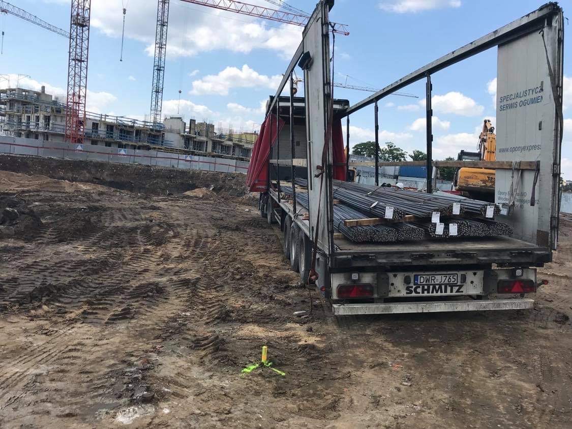 Ciężarówka z rozsuwanym dachem i bokami, załadowana stalowymi prętami, stoi na błotnistym placu budowy z widocznymi dźwigami i powstającym budynkiem w tle.