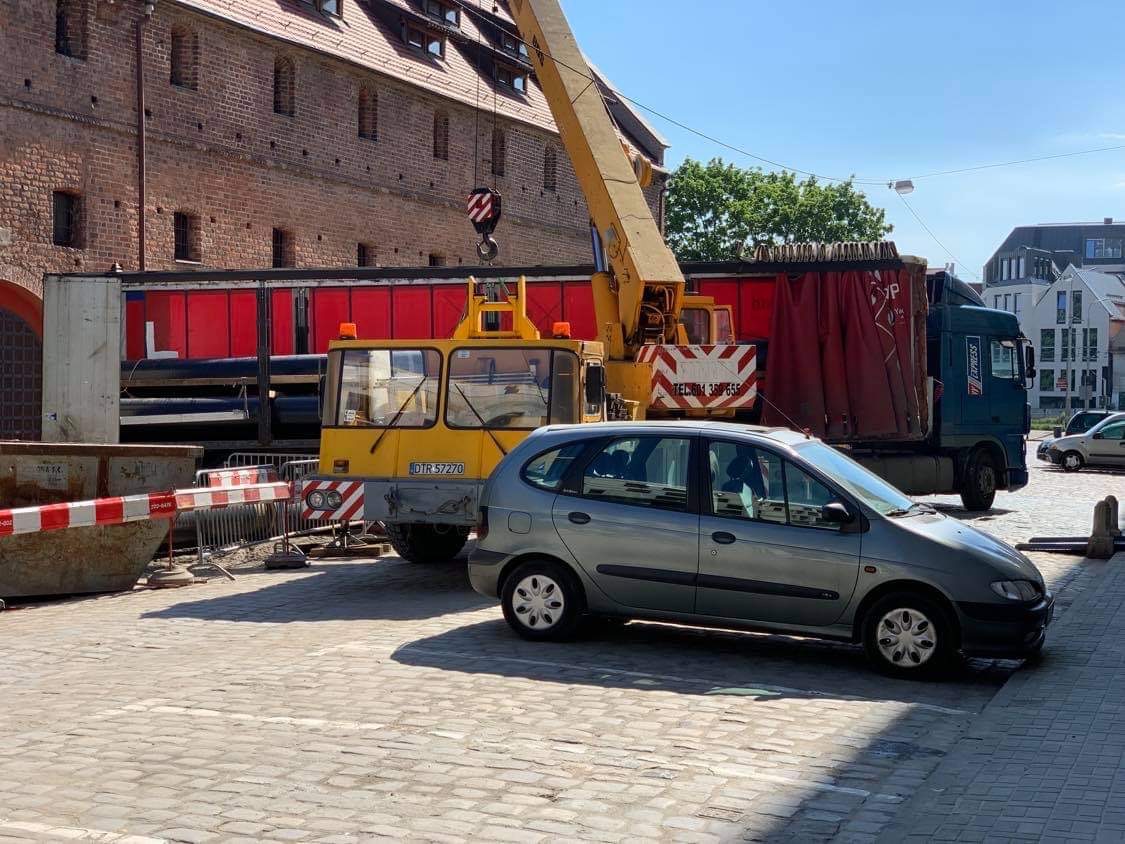 Żółty dźwig mobilny z czerwoną liną i hakiem, zaparkowany obok czerwonej ciężarówki z czarnymi rurami na tle ceglanego budynku, z szarym samochodem osobowym na pierwszym planie.