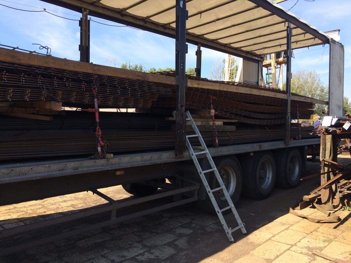 Ciężarówka z odkrytą naczepą załadowana stalowymi prętami i siatkami, zabezpieczonymi czerwonymi pasami, z aluminiową drabiną opartą o bok naczepy.