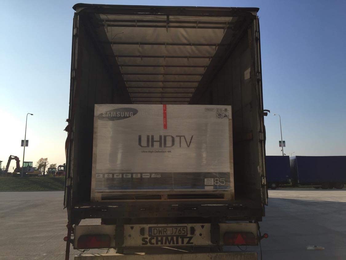 Paleta z zapakowanym telewizorem Samsung UHDTV 85 cali wewnątrz naczepy ciężarówki Schmitz na parkingu, w tle widoczny sprzęt budowlany i inne ciężarówki.