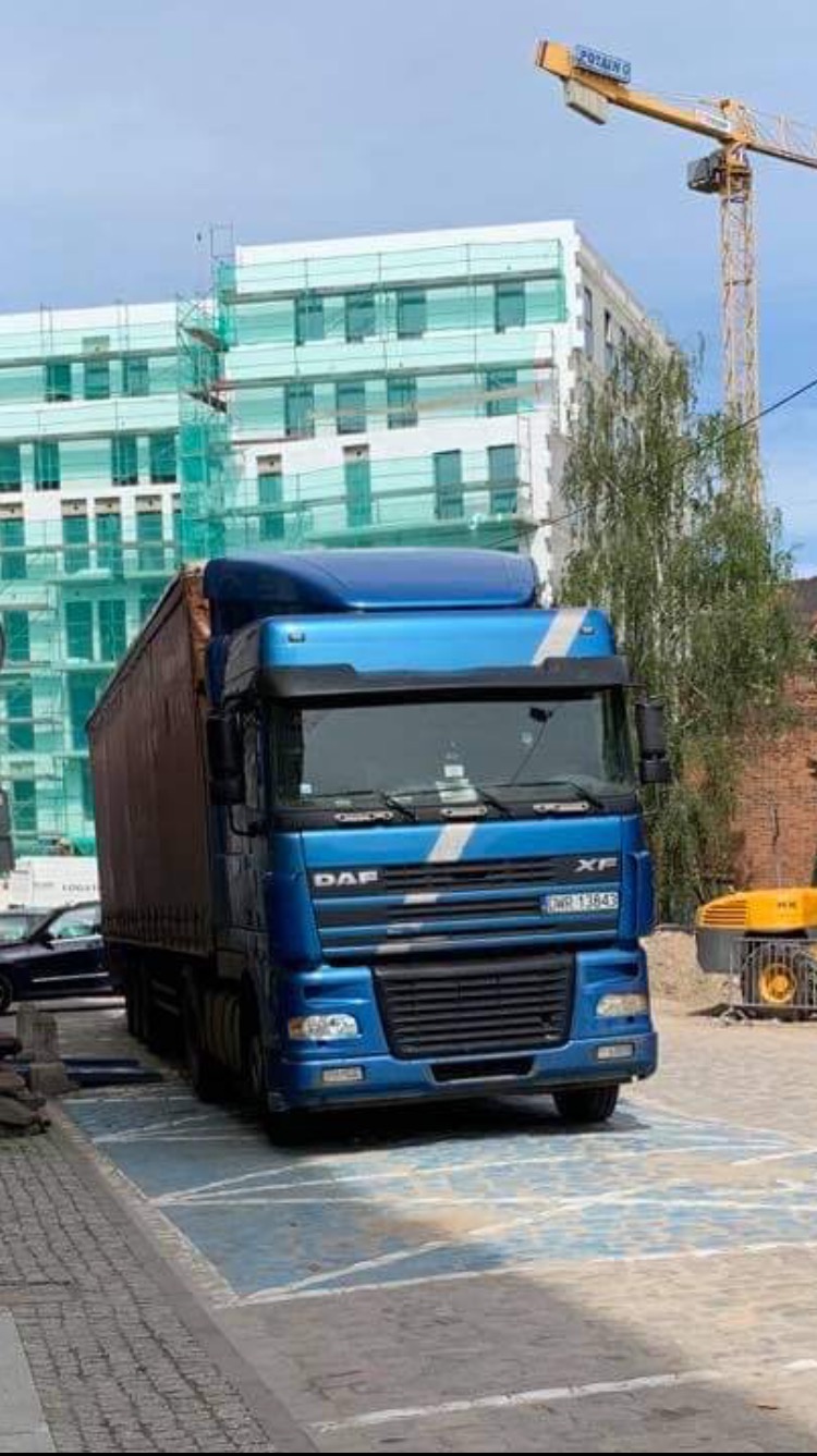 Niebieski ciągnik siodłowy DAF XF z naczepą zaparkowany na wyznaczonym miejscu parkingowym, w tle budynek w rusztowaniach i żuraw budowlany.