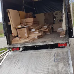 Wnętrze samochodu dostawczego wypełnione kartonami i meblami gotowymi do transportu, widok z tyłu na podniesioną rampę załadunkową.