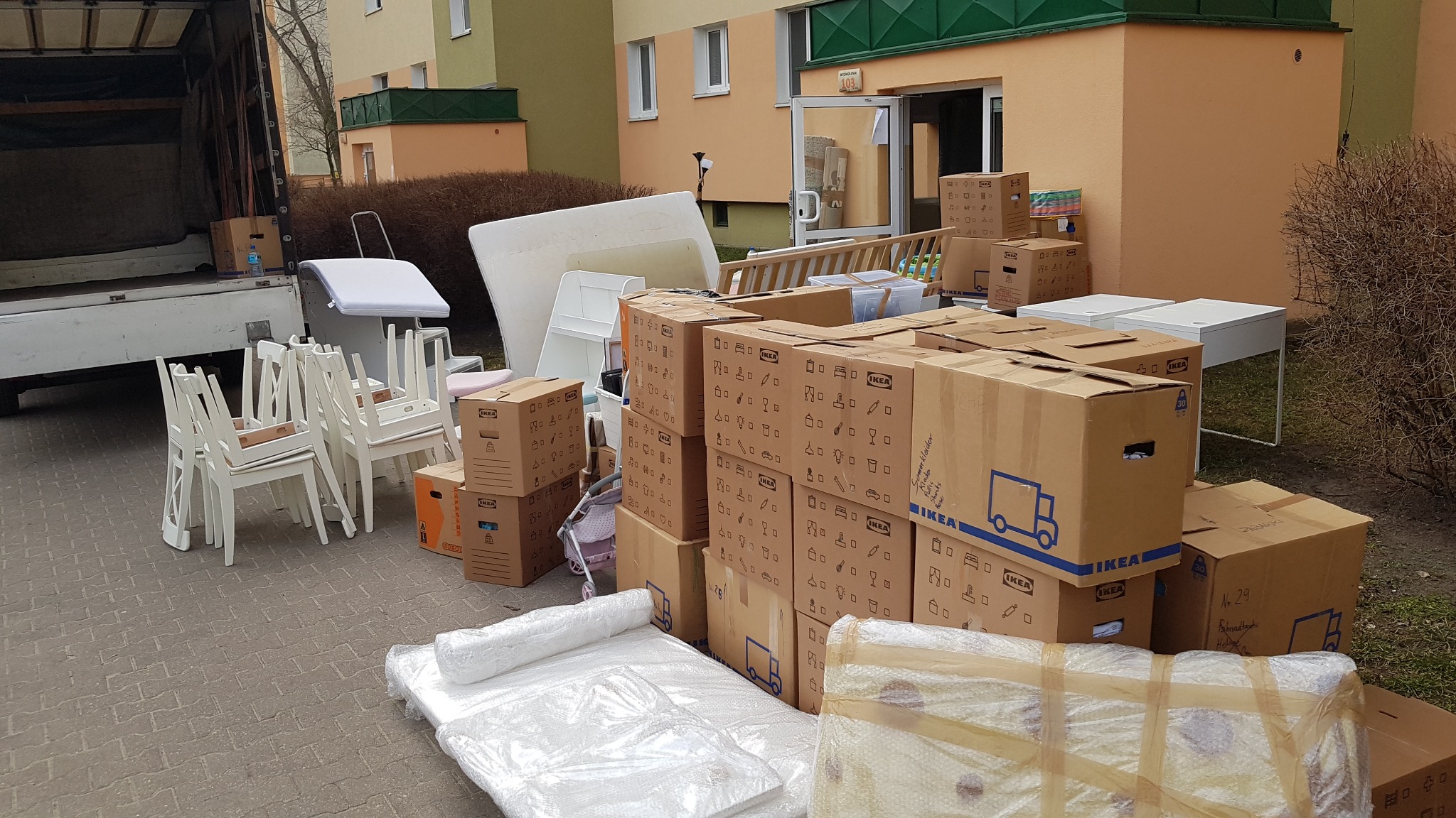 Przygotowania do przeprowadzki: kartony IKEA, meble owinięte folią, białe krzesła i materac przed budynkiem mieszkalnym, częściowo załadowane do ciężarówki.