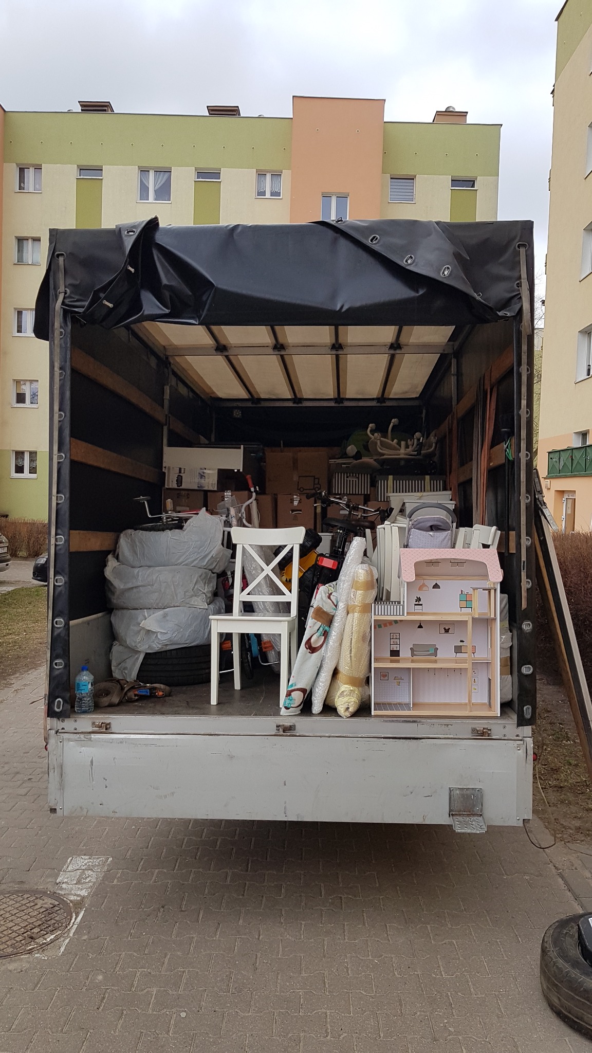 Widok z tyłu na załadowaną ciężarówkę z plandeką, wypełnioną meblami, kartonami i przedmiotami gospodarstwa domowego, gotową do transportu podczas przeprowadzki.