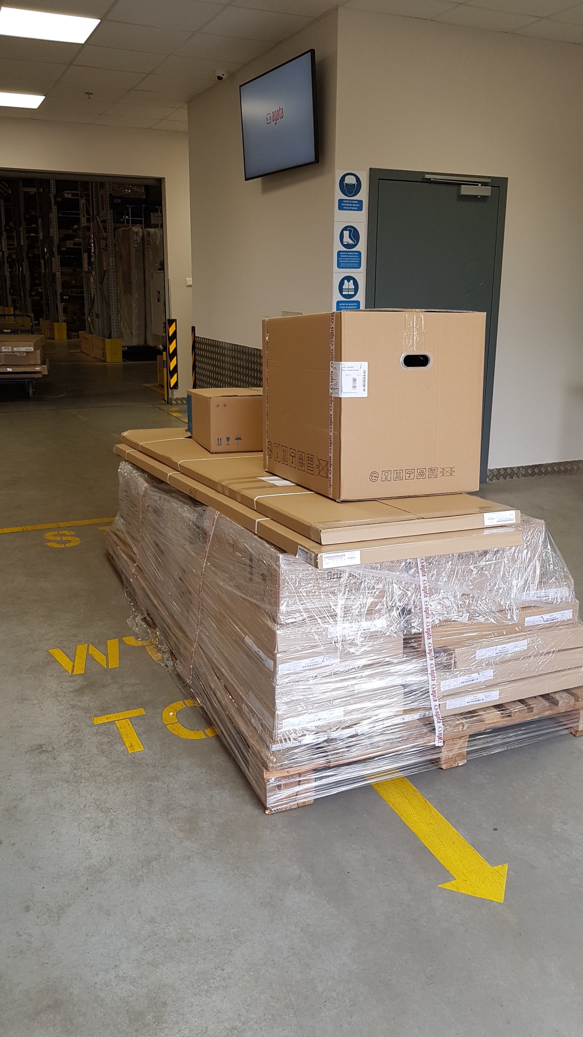 Paleta z zapakowanymi kartonami i elementami meblowymi, owinięta folią stretch, w magazynie z żółtymi oznaczeniami na podłodze.