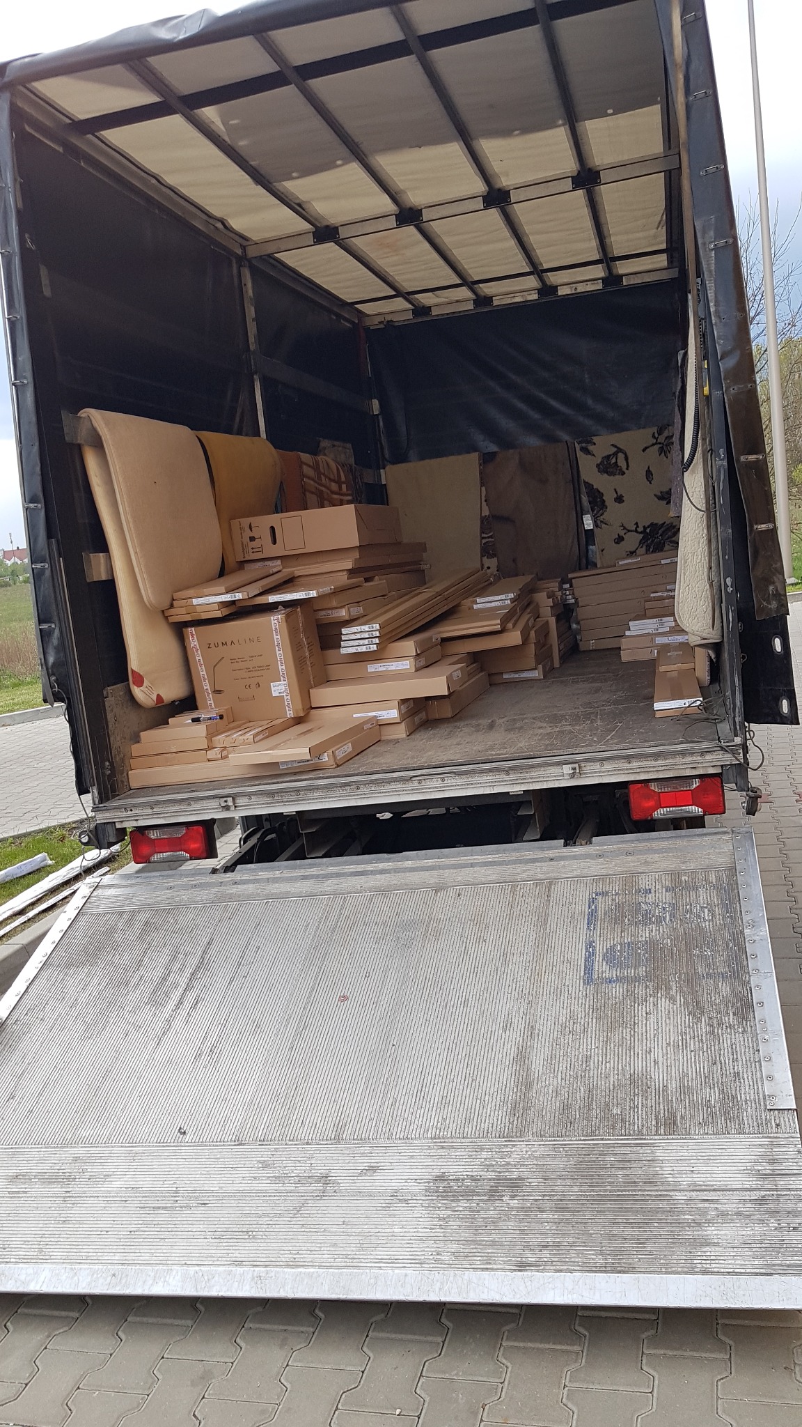 Wnętrze samochodu dostawczego wypełnione kartonami i meblami gotowymi do transportu, widok z tyłu na podniesioną rampę załadunkową.