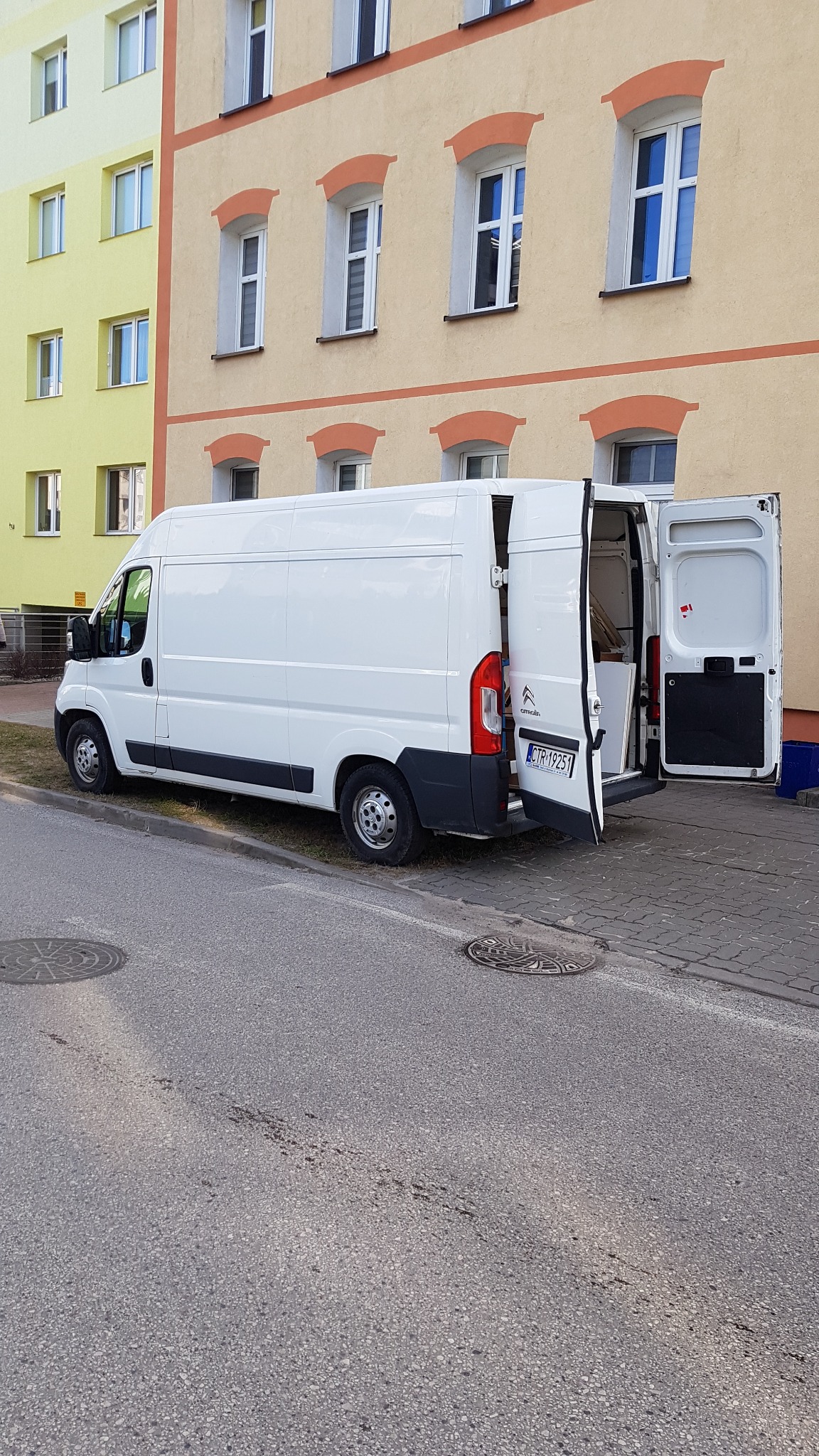 Biały furgon Citroen z otwartymi tylnymi drzwiami zaparkowany na ulicy przed budynkiem mieszkalnym o żółto-pomarańczowej fasadzie.
