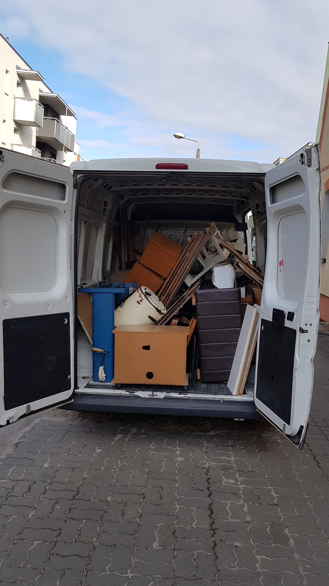 Otwarty bagażnik białego busa wypełniony różnymi meblami i przedmiotami podczas przeprowadzki na brukowanym podwórku, w tle budynek mieszkalny.
