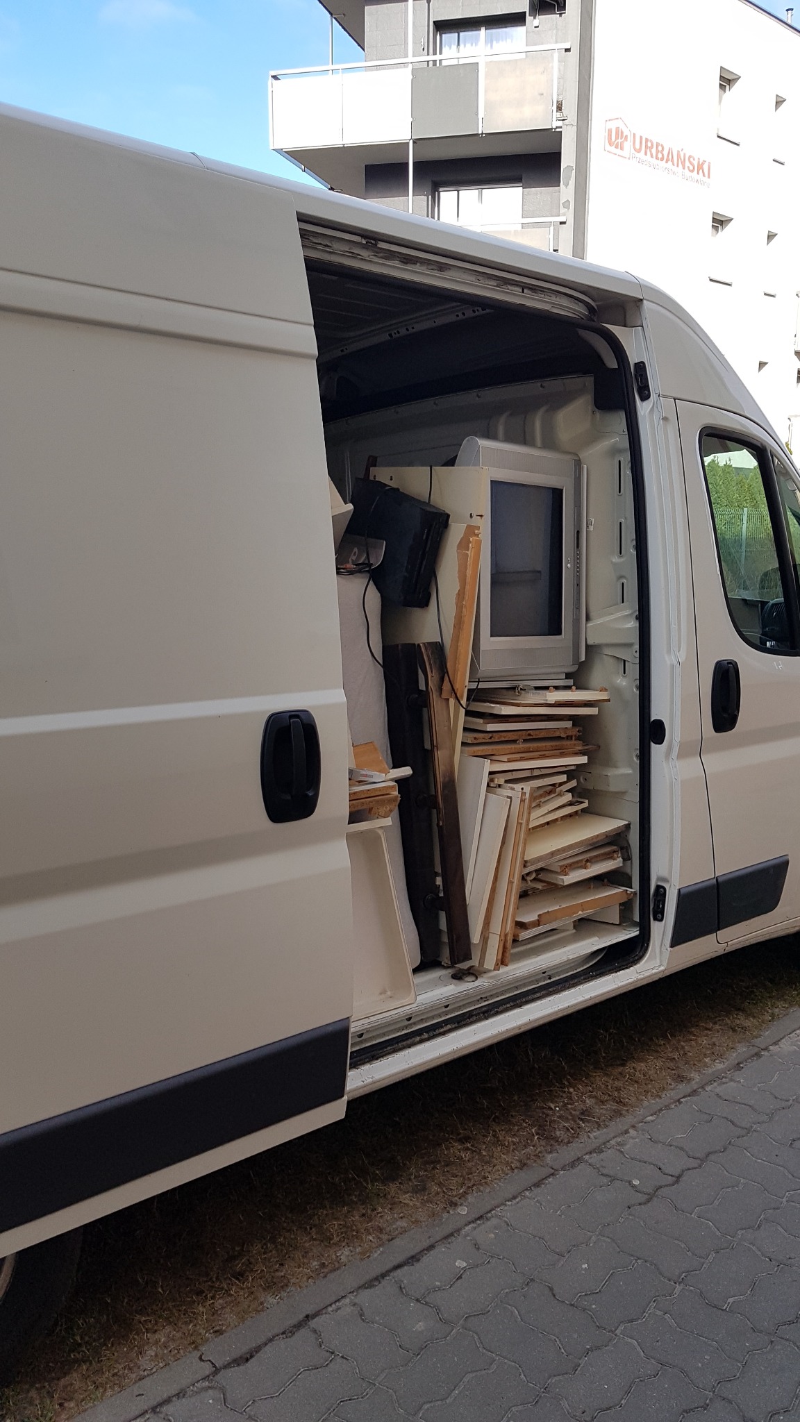 Otwarty biały furgon wypełniony kartonami, meblami i starym telewizorem kineskopowym podczas transportu na tle budynku mieszkalnego z logo firmy budowlanej.