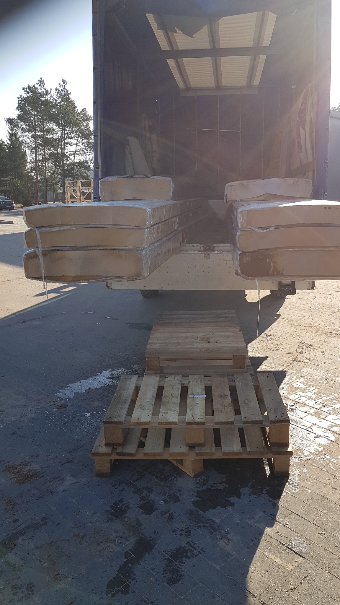 Załadunek spakowanych materacy z ciężarówki na palety, widok z tyłu ciężarówki, słoneczny dzień.