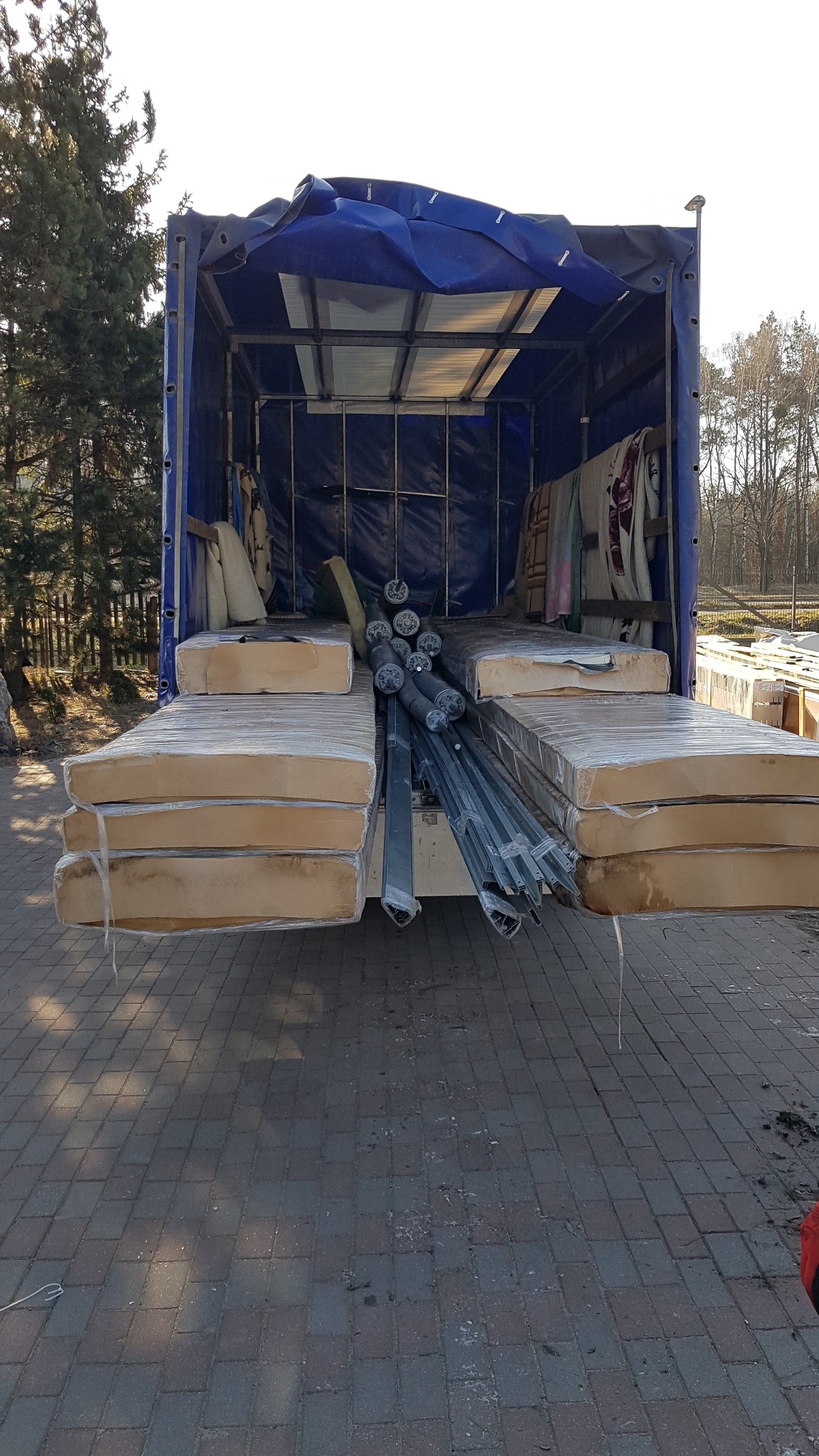 Otwarta naczepa ciężarówki z niebieską plandeką załadowana materacami w folii, metalowymi profilami i innymi przedmiotami podczas dnia.