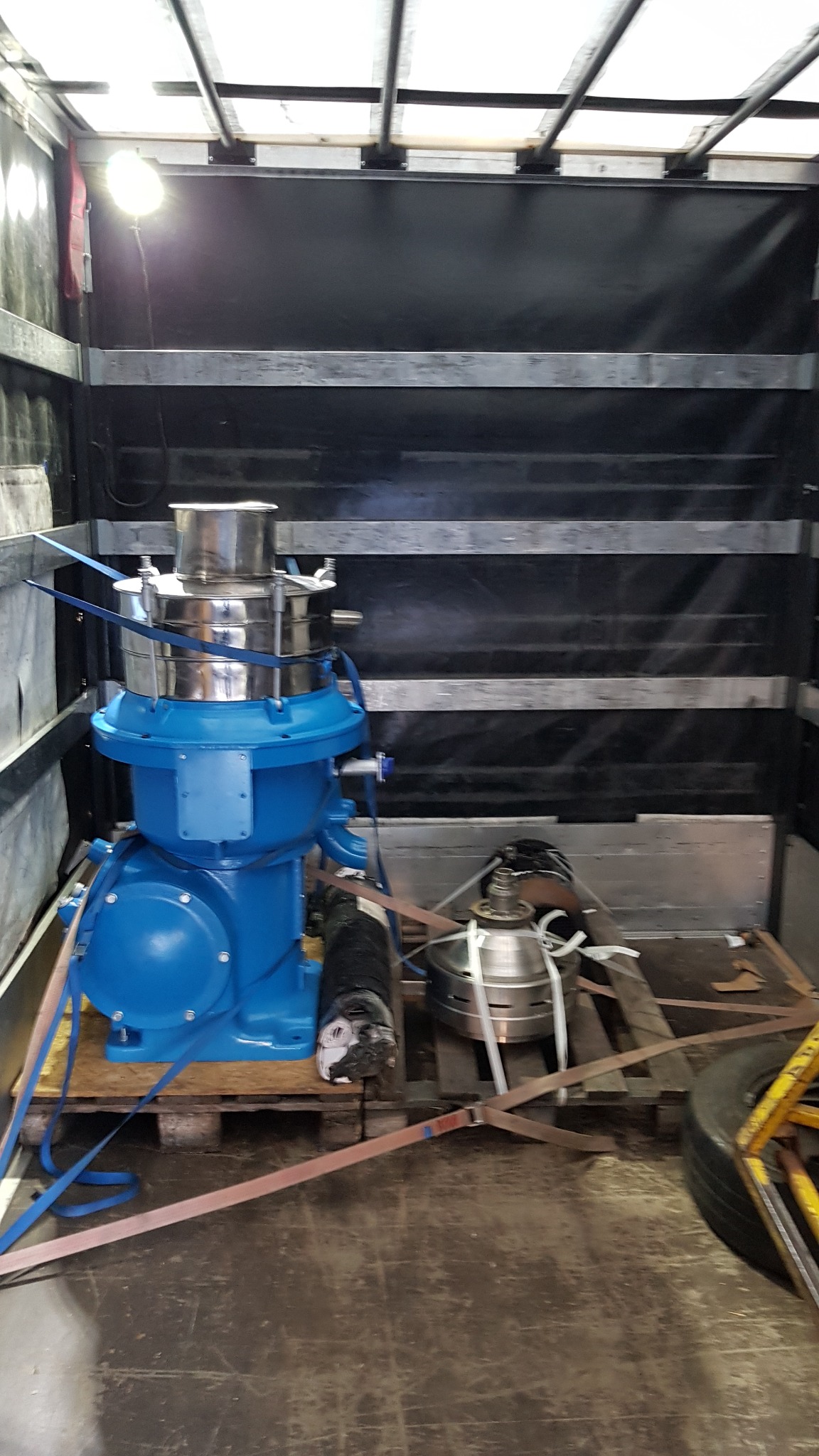 Niebieska maszyna przemysłowa zabezpieczona pasami transportowymi na palecie wewnątrz ciemnej furgonetki z oświetleniem sufitowym.