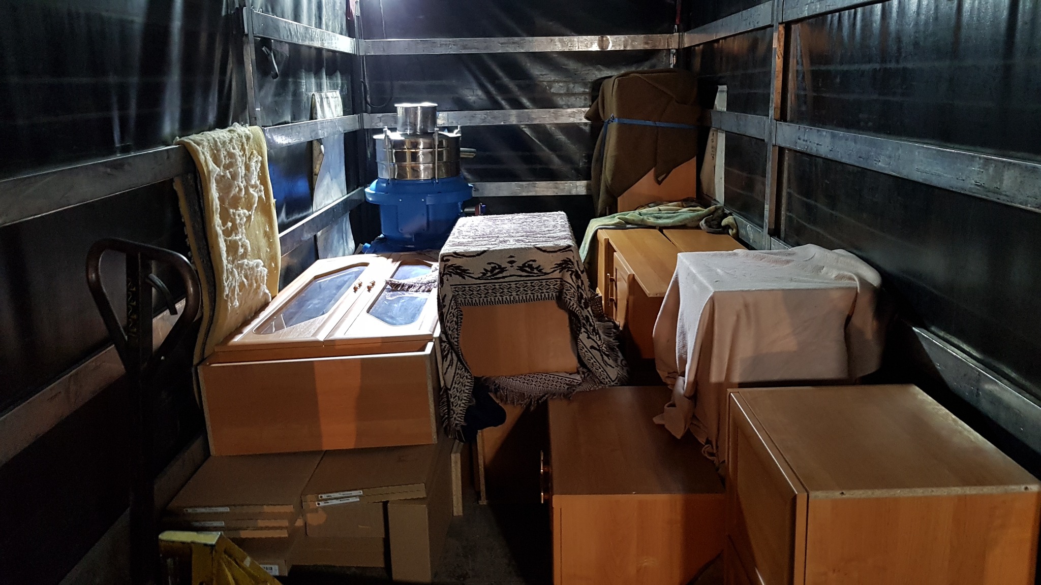 Wnętrze samochodu dostawczego wypełnione meblami i sprzętem, przygotowanymi do transportu, z widocznym niebieskim urządzeniem przemysłowym pośród zapakowanych szafek i komód.