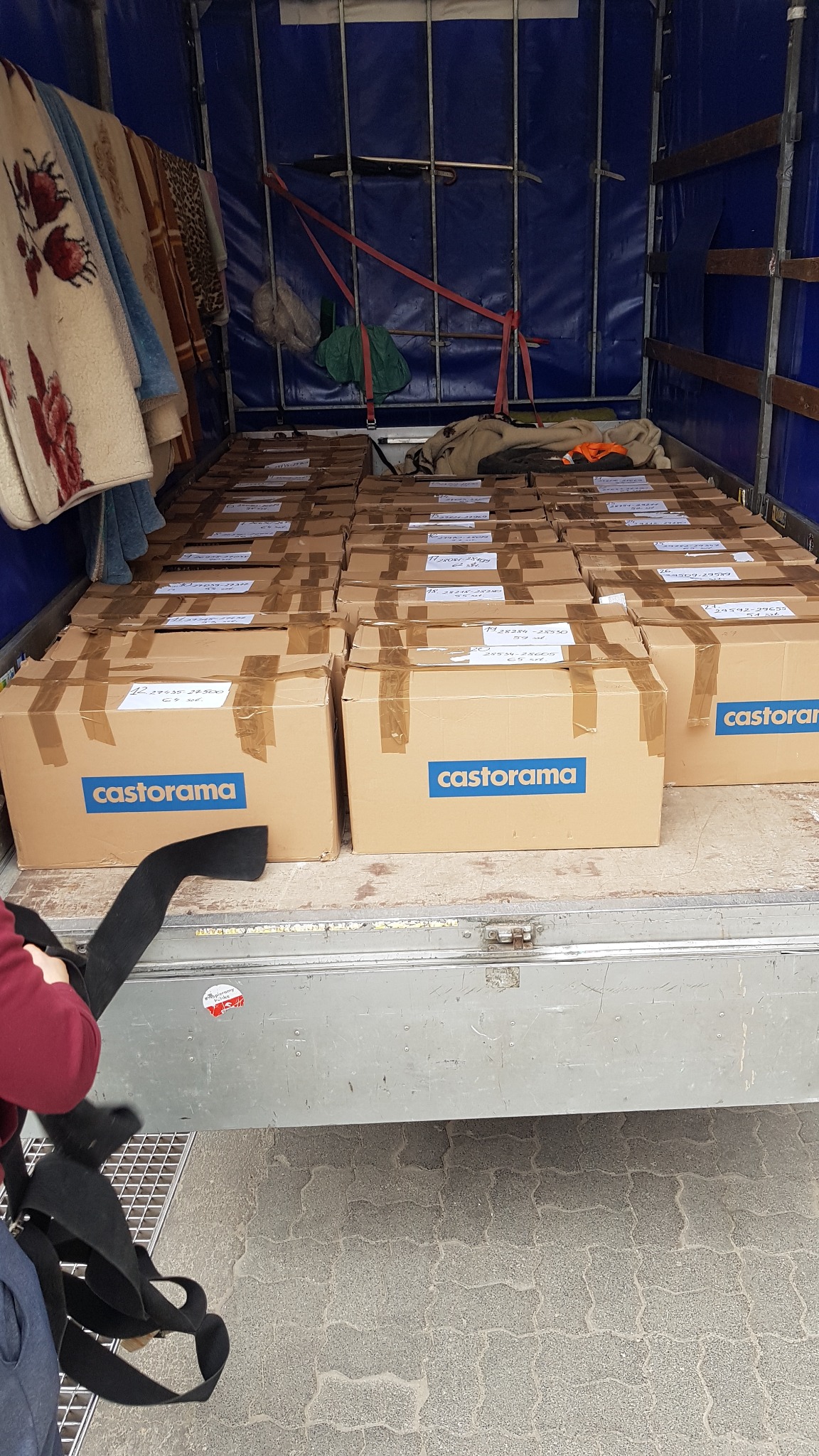 Wnętrze ciężarówki wypełnione kartonami z logo Castorama, zabezpieczonymi taśmą, gotowymi do transportu; w tle widoczne koce i ubrania, sugerujące przeprowadzkę lub transport towarów.