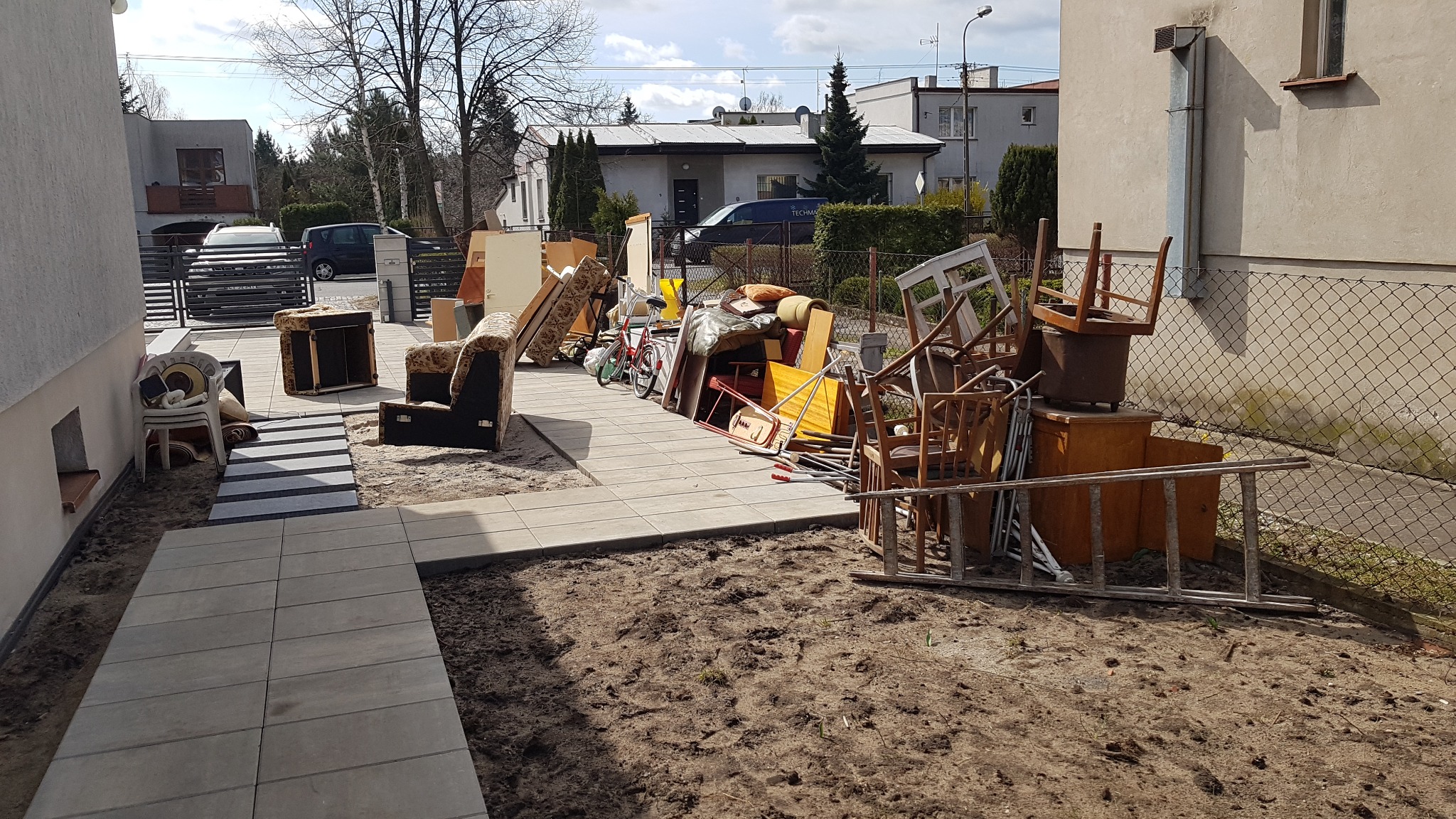 Stos mebli i przedmiotów przygotowanych do wyniesienia przed domem, widoczny fragment chodnika z szarej kostki.