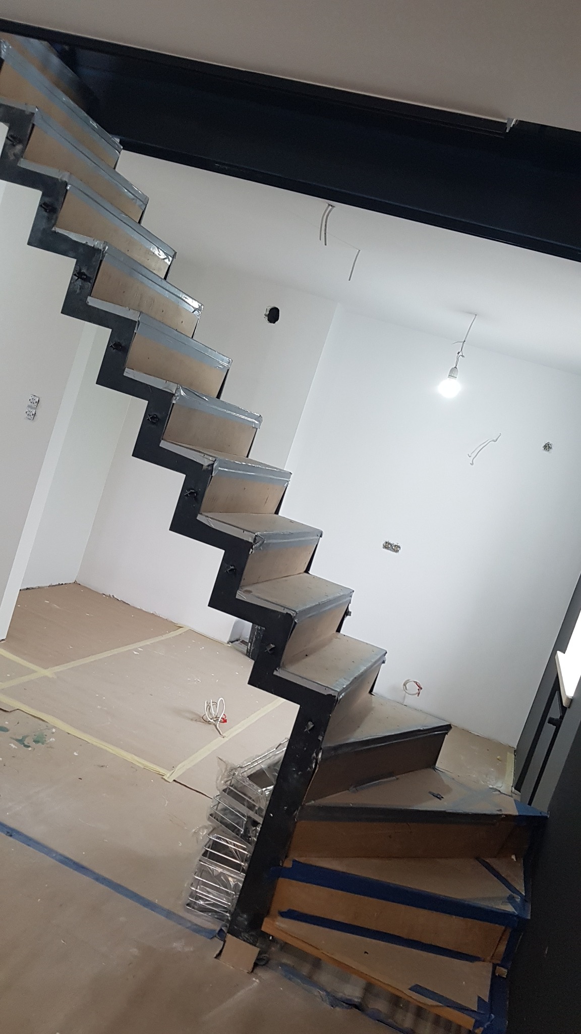 Nowoczesne schody ażurowe z czarną konstrukcją stalową i drewnianymi stopniami, zabezpieczone folią ochronną podczas remontu, widoczne przygotowania do montażu balustrady.