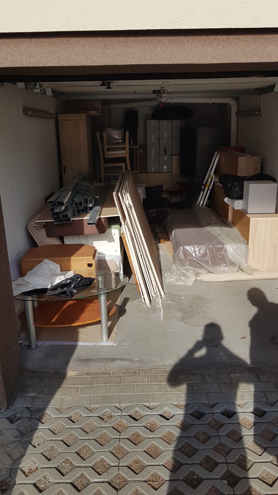 Garaż wypełniony meblami i kartonami przygotowanymi do transportu, widoczne elementy szklanego stolika, płyty meblowe i metalowe profile, zarys sylwetki osoby w dolnej części kadru.