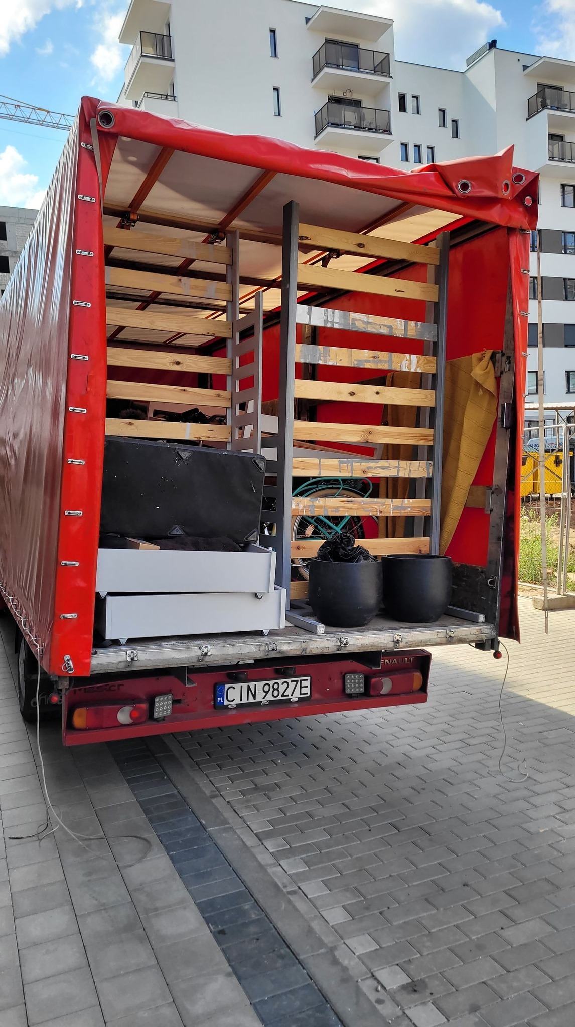 Otwarta skrzynia ładunkowa czerwonej ciężarówki wypełniona meblami, w tle nowoczesny budynek mieszkalny.