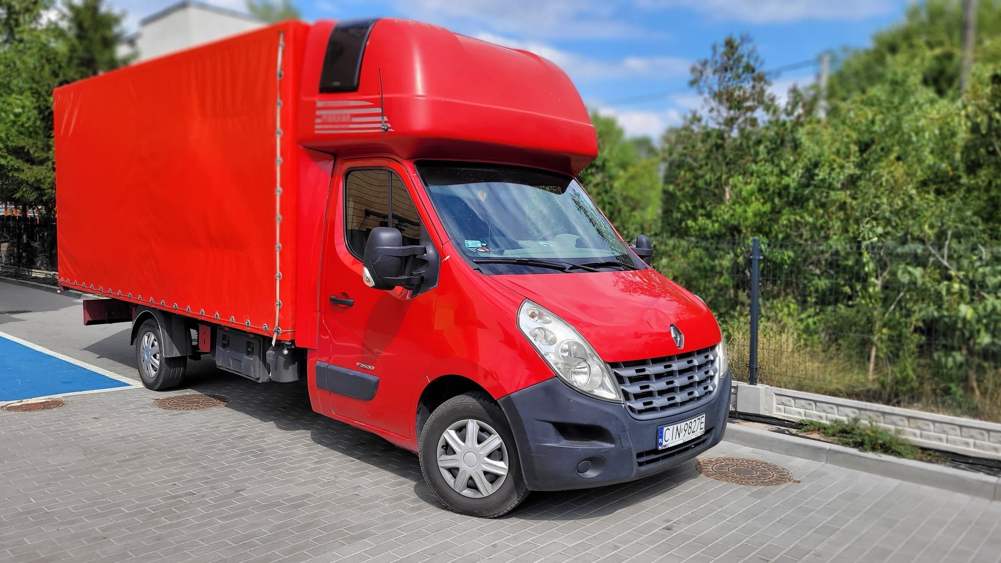 Czerwony samochód dostawczy Renault Master z plandeką, zaparkowany na kostce brukowej, z widoczną tablicą rejestracyjną i zielenią w tle.