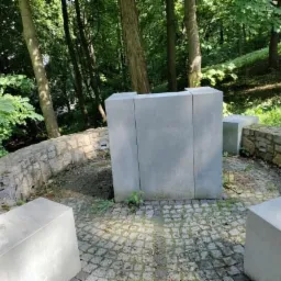 Betonowa instalacja artystyczna na brukowanym placu w otoczeniu drzew, z elementami kamiennej architektury w tle, prawdopodobnie na Zamku w Będzinie.