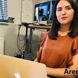 Kobieta z lekkim uśmiechem pracuje na laptopie, w tle biurko z dwoma monitorami i jasne pomieszczenie biurowe, widoczne logo 'Arena' w prawym dolnym rogu.