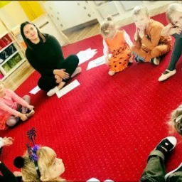 Grupa dzieci siedzi w kręgu na czerwonym dywanie podczas lekcji z nauczycielką, białe kartki porozrzucane na podłodze, regał z zabawkami w tle.