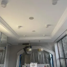 Biały sufit z zamontowanymi okrągłymi lampami punktowymi oraz listwami dekoracyjnymi, widoczne lampy wiszące zabezpieczone folią malarską, ślady prac remontowych.