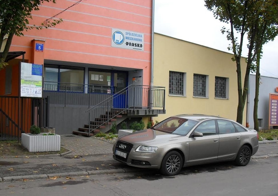 Srebrny samochód Audi zaparkowany przed budynkiem Spółdzielni Mieszkaniowej Piaski z widoczną tablicą informacyjną dotyczącą ubezpieczeń.