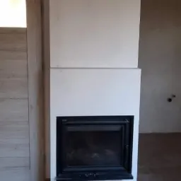 Wnętrze z kominkiem w trakcie wykończenia, widoczne płytki podłogowe imitujące drewno i niedokończone ściany.