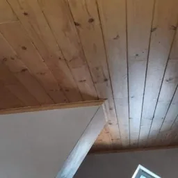 Wykończony drewniany sufit z widocznymi sękami i lampami wiszącymi, krawędź ściany pomalowana na biało, widok z dołu.