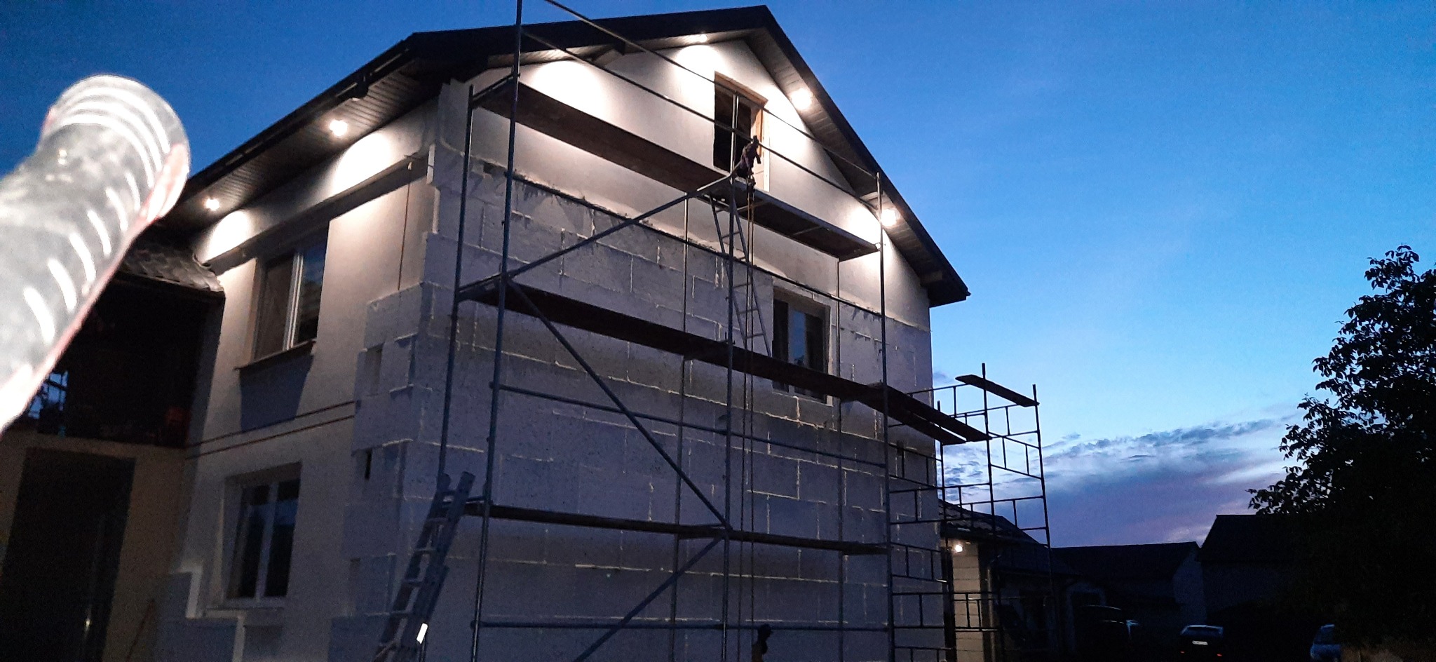 Dom w trakcie ocieplania elewacji zewnętrznej, widoczne rusztowania i warstwa izolacyjna, oświetlenie elewacji włączone, wieczorny krajobraz.