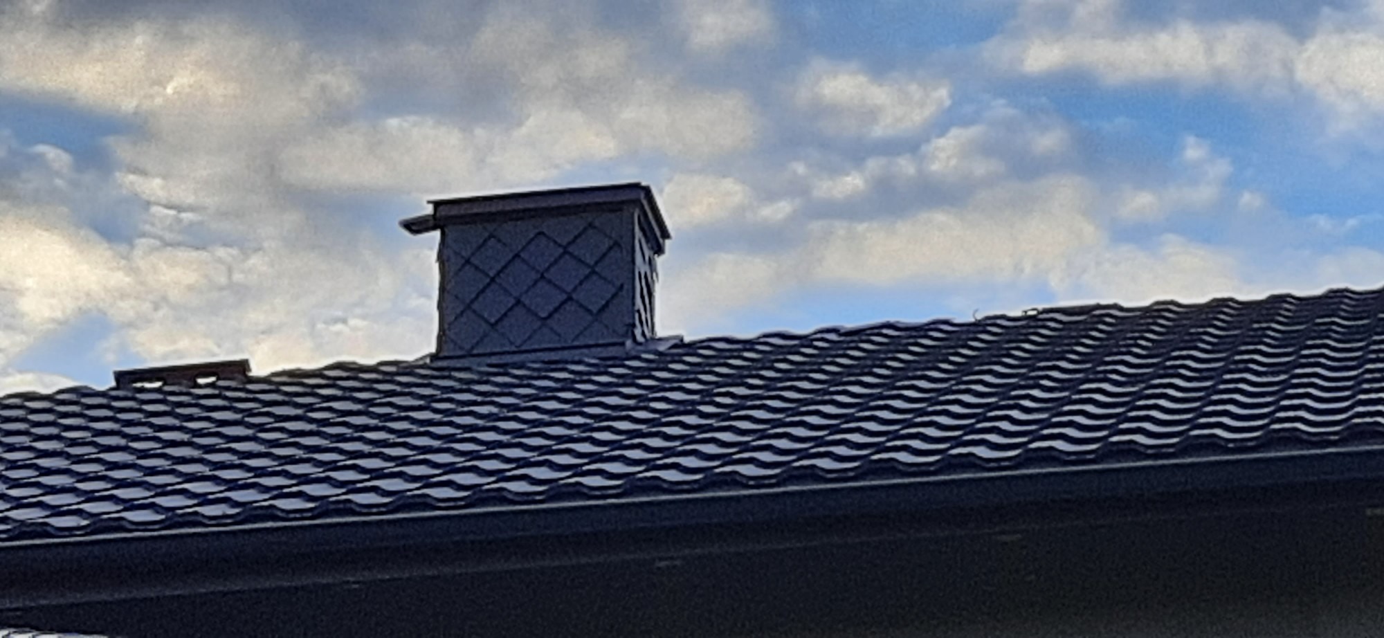 Nowy, ciemny dach z blachy trapezowej i komin z dekoracyjną obudową na tle pochmurnego nieba.