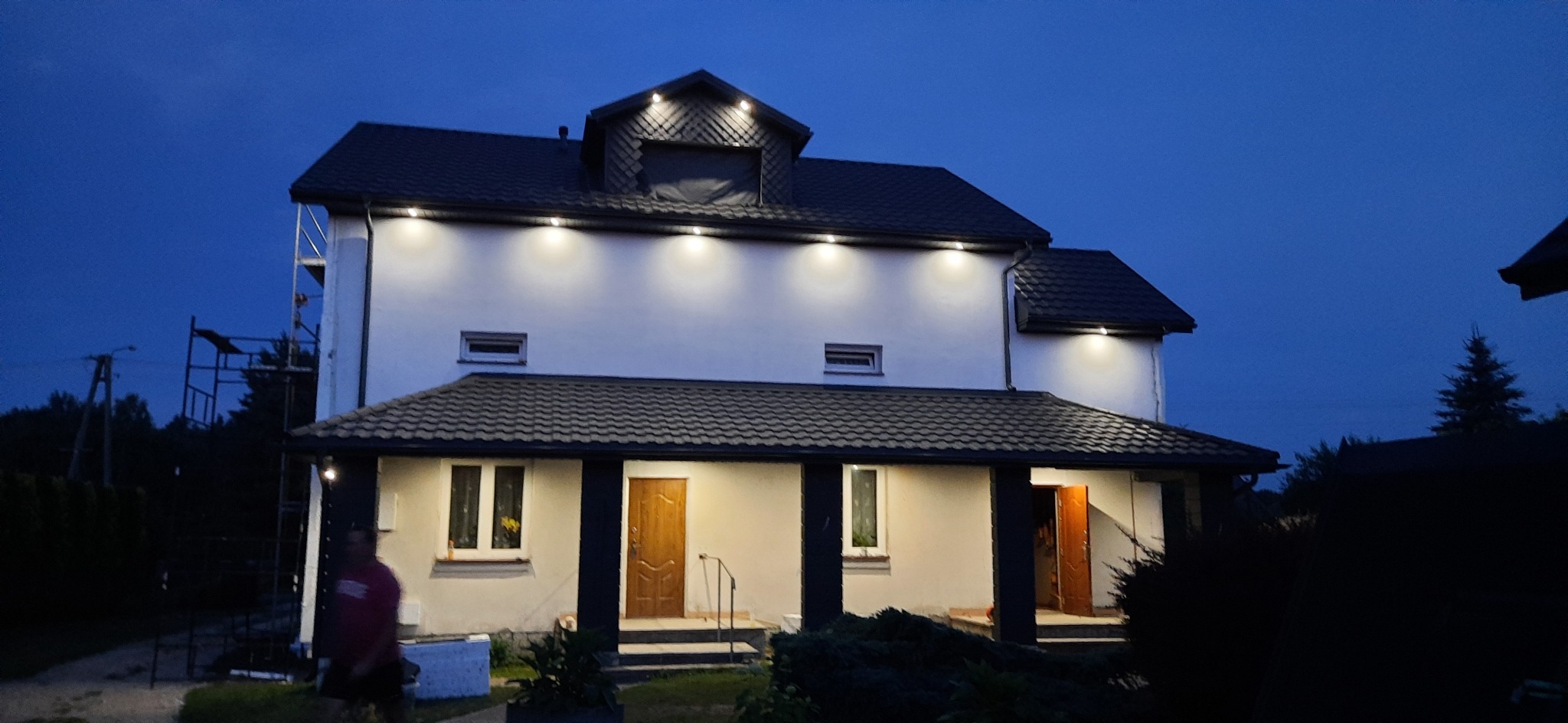 Dwupiętrowy dom z szarą dachówką i białymi ścianami oświetlony zewnętrznymi lampami w nocnej scenerii; widoczne rusztowanie z boku budynku.