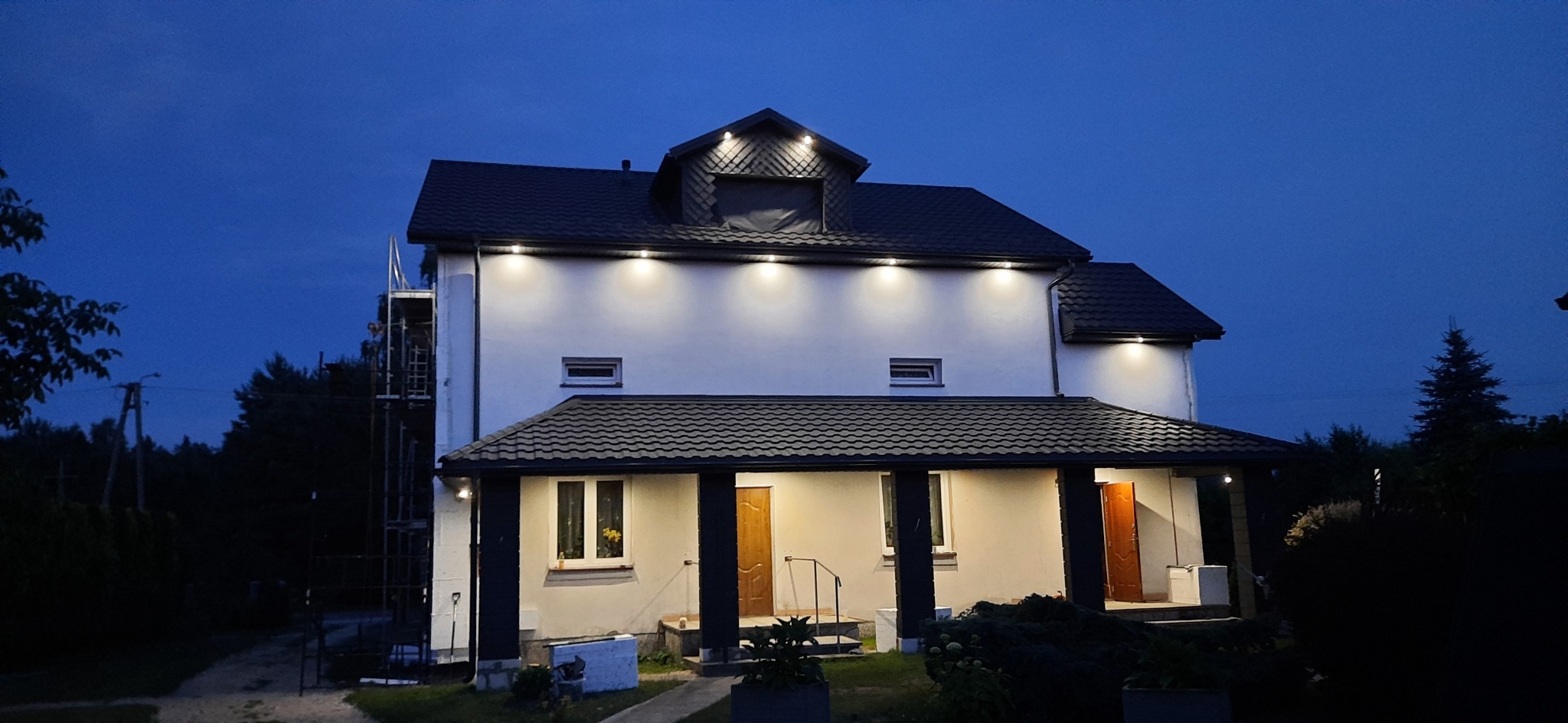 Dwupiętrowy dom z oświetloną elewacją i nowym dachem, widziany o zmierzchu, z rusztowaniem z boku budynku.