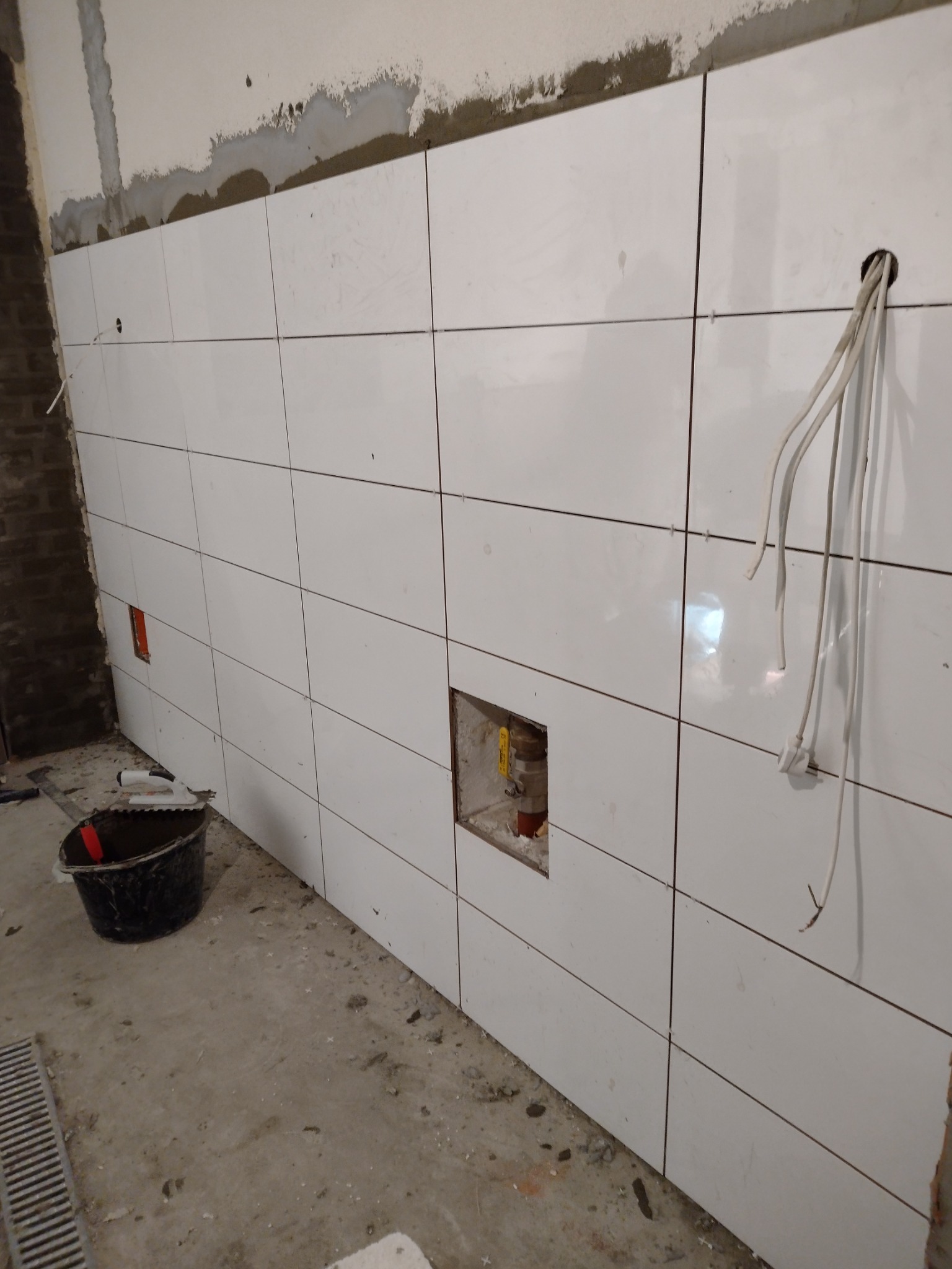 Białe płytki ceramiczne układane na ścianie w łazience, widoczne otwory na instalacje, wiadro z zaprawą i wystające przewody elektryczne.