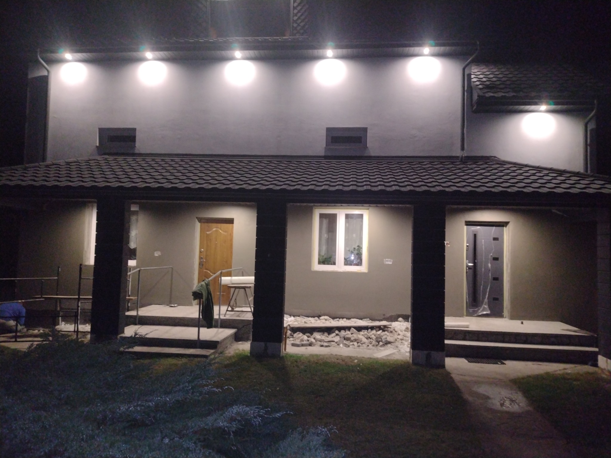 Nocne ujęcie oświetlonego domu z szarą elewacją, widocznymi nowymi drzwiami wejściowymi, świeżo położoną dachówką i przygotowanym terenem pod elewacją werandy.