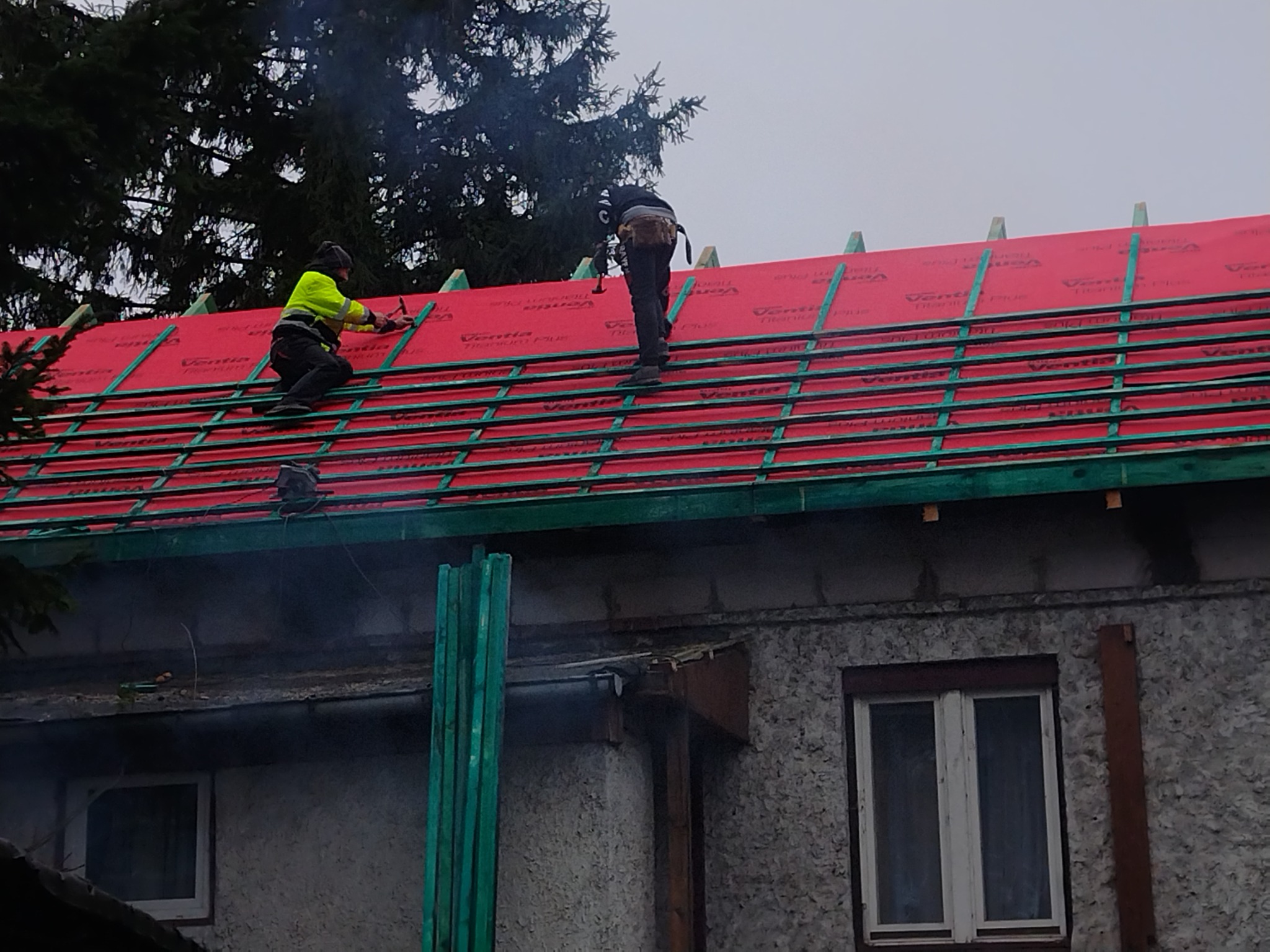 Dwóch dekarzy pracuje na dachu domu, mocując czerwone membrany dachowe do zielonej konstrukcji drewnianej pod spodem. Jeden dekarz ubrany w żółtą kurtkę wbija gwoździe młotkiem, drugi stoi obok.
