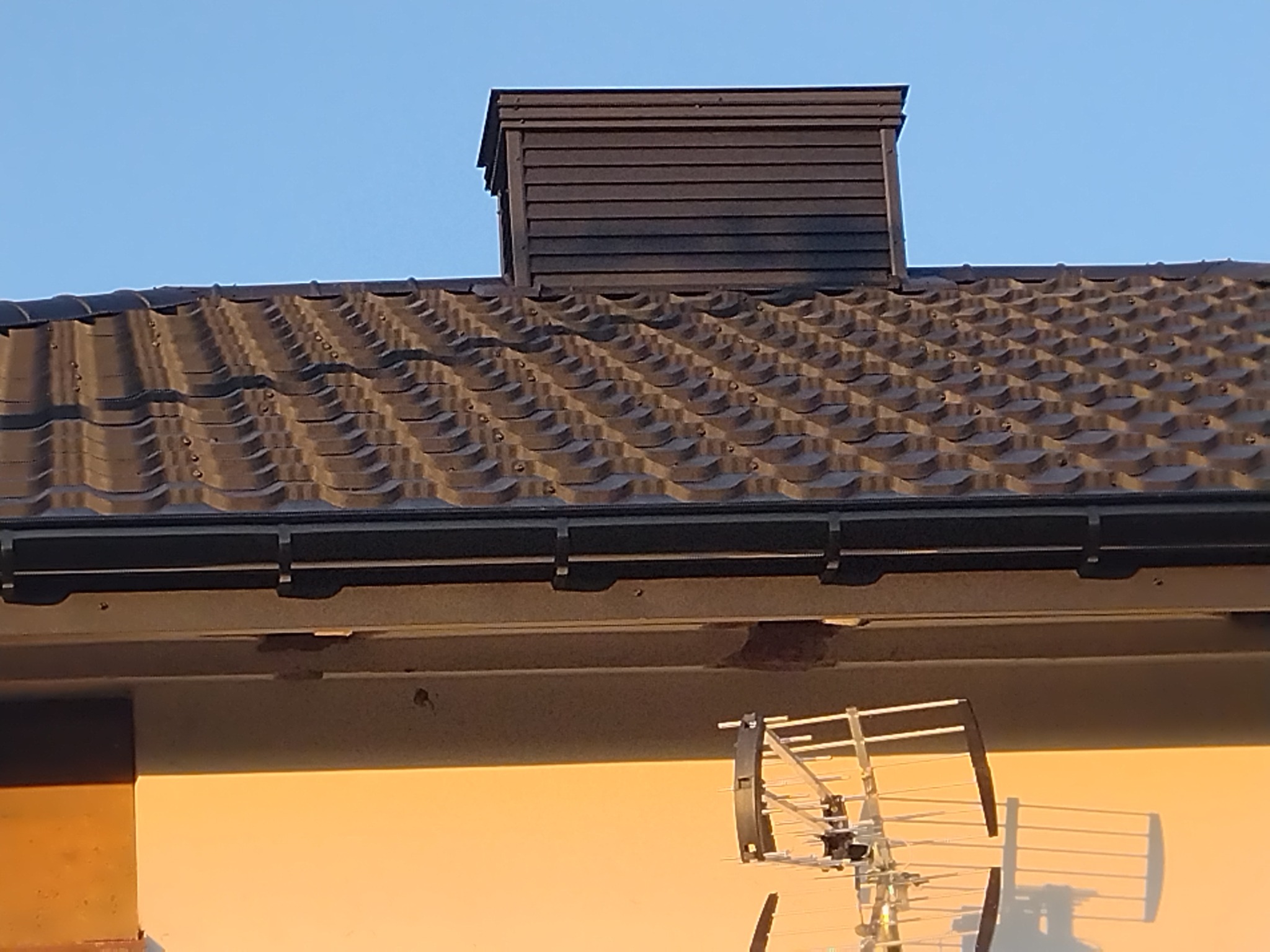 Ciemna dachówka ceramiczna z kominem wentylacyjnym na nowym dachu, widoczne orynnowanie i fragment ściany budynku z anteną telewizyjną.