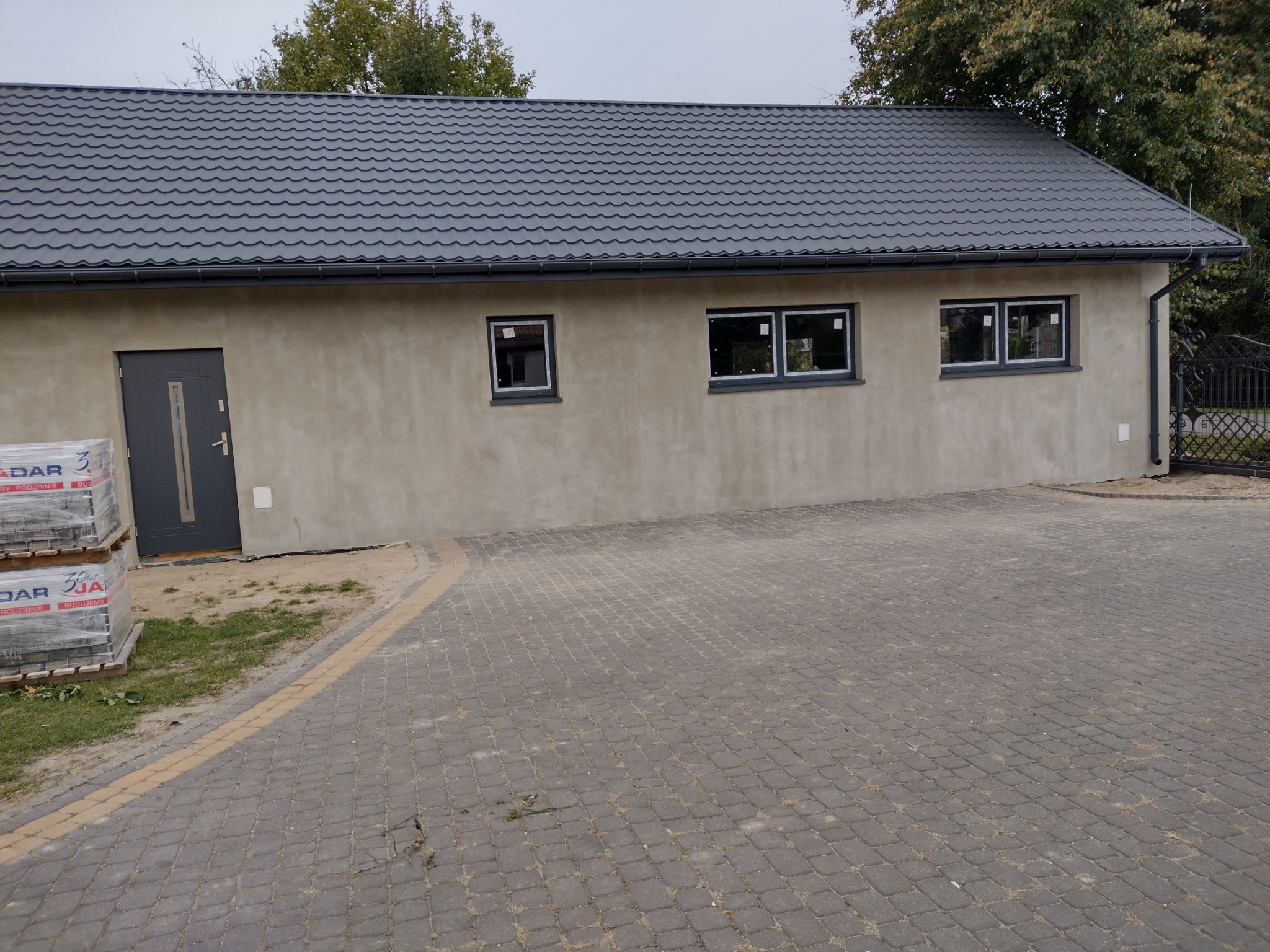 Nowy budynek z szarą blachodachówką, trzema oknami z ciemnymi ramami, szarymi drzwiami i stosem materiałów budowlanych przed wejściem.