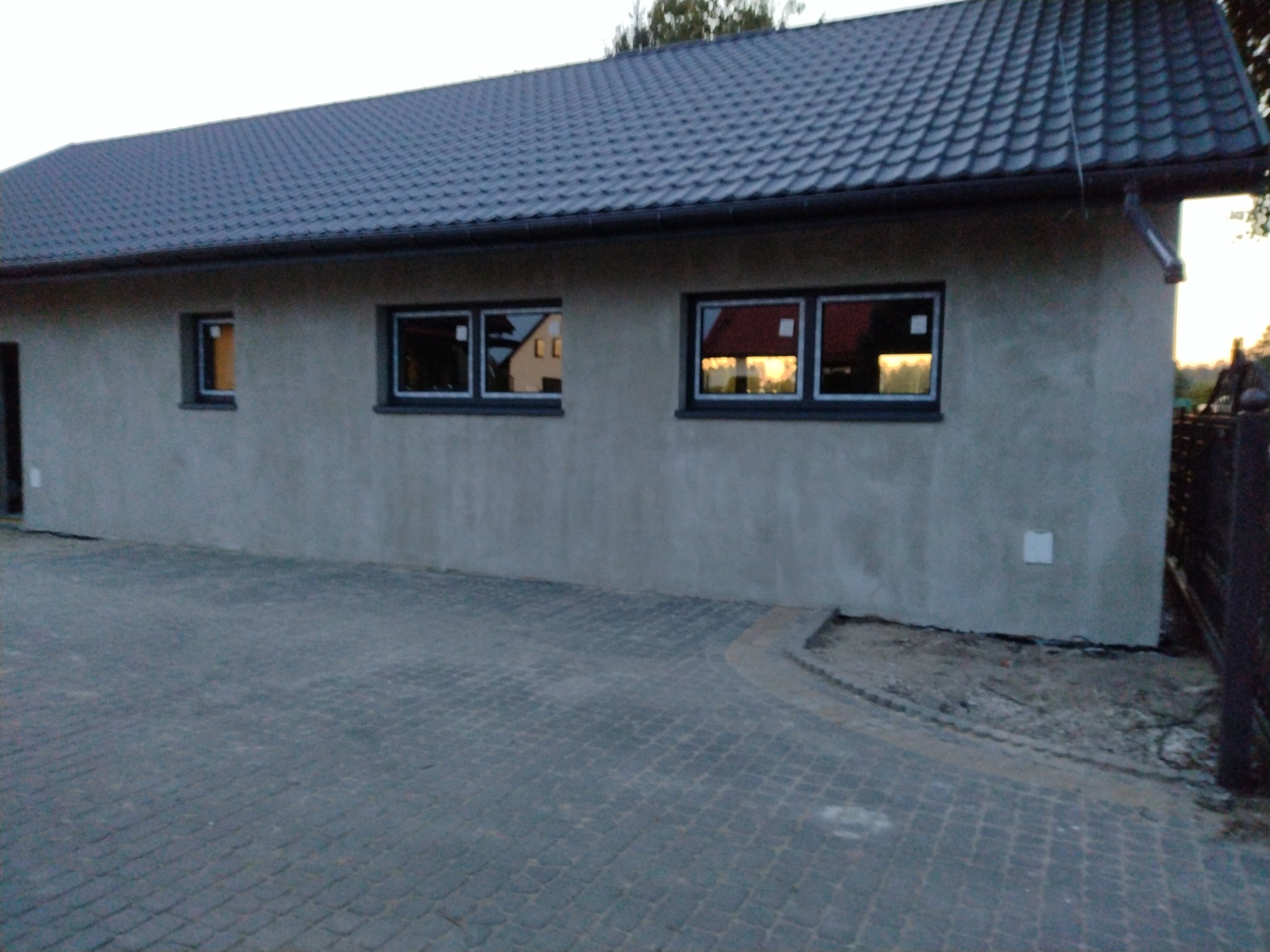 Budynek po remoncie: nowa, ciemnoszara dachówka, szara, otynkowana elewacja z trzema oknami w ciemnych ramach. W oknach odbija się zachodzące słońce i sąsiedni budynek. Podjazd wyłożony kostką...