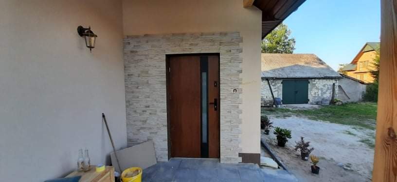 Drewniane drzwi wejściowe z czarnymi akcentami, otoczone dekoracyjną okładziną z kamienia elewacyjnego, zamontowane na zewnątrz budynku z widocznym oświetleniem i elementami otoczenia.