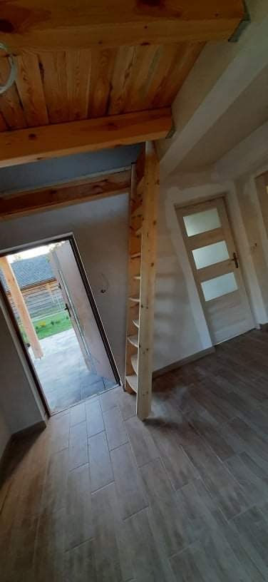 Wnętrze domu w trakcie wykańczania: drewniany sufit, drzwi wewnętrzne z przeszkleniami, podłoga wyłożona płytkami imitującymi drewno. Widoczna konstrukcja schodów i otwarte drzwi na zewnątrz...