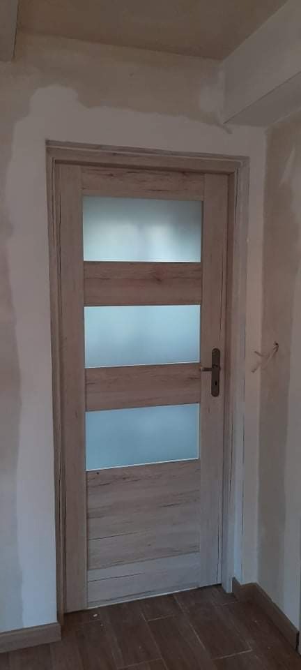 Zamknięte, drewnopodobne drzwi wewnętrzne z trzema poziomymi szybami z mlecznego szkła, zamontowane w surowym otworze, widoczne ślady po pracach wykończeniowych na ścianach i suficie.
