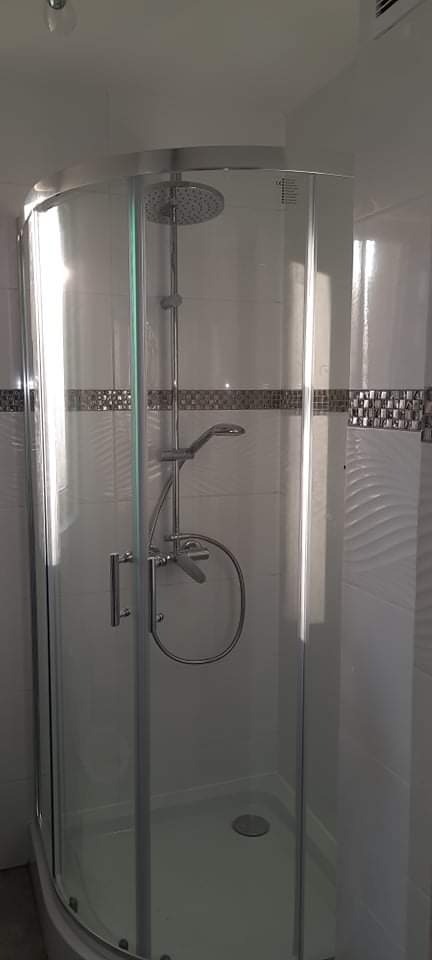 Półokrągła kabina prysznicowa z chromowanymi elementami i deszczownicą, białe płytki z dekoracyjnym pasem mozaiki, widok na wykończoną łazienkę.