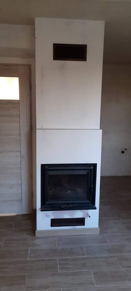 Wnętrze z kominkiem w trakcie wykończenia, widoczne płytki podłogowe imitujące drewno i niedokończone ściany.