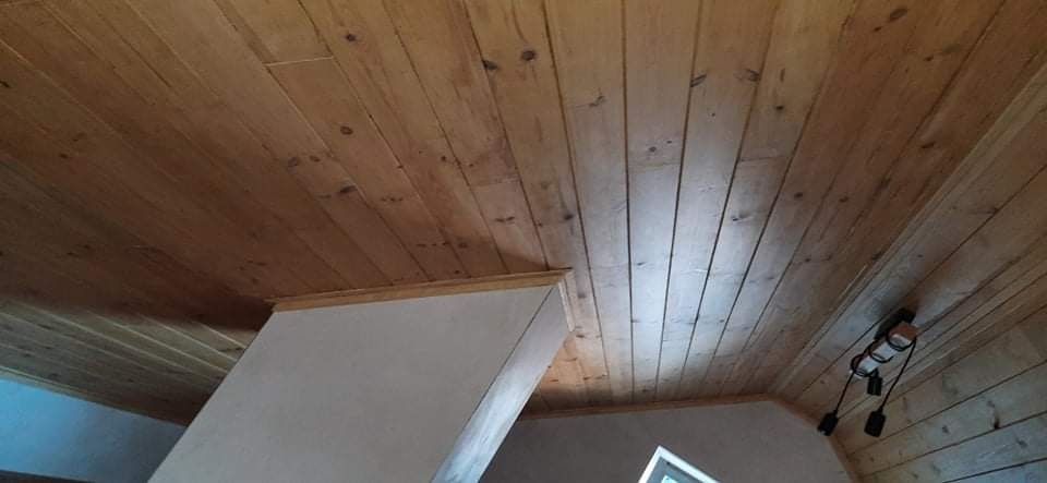 Wykończony drewniany sufit z widocznymi sękami i lampami wiszącymi, krawędź ściany pomalowana na biało, widok z dołu.