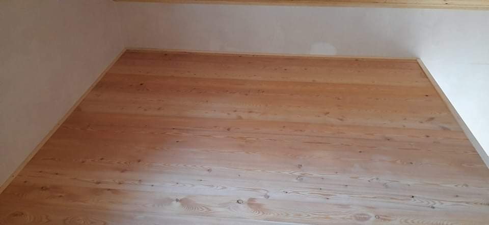 Jasny, drewniany parkiet z widocznymi słojami, ułożony w prostym wzorze w rogu pokoju z białymi ścianami i drewnianą listwą przypodłogową.