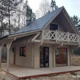 Gotowy, parterowy dom z bali z ciemną dachówką, drewnianym balkonem z ozdobnymi krzyżami i zadaszonym tarasem, otoczony lasem.