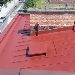 Czerwony, metalowy dach z kominem, anteną satelitarną i panelami słonecznymi widziany z góry, z widokiem na ulicę i budynki w tle.